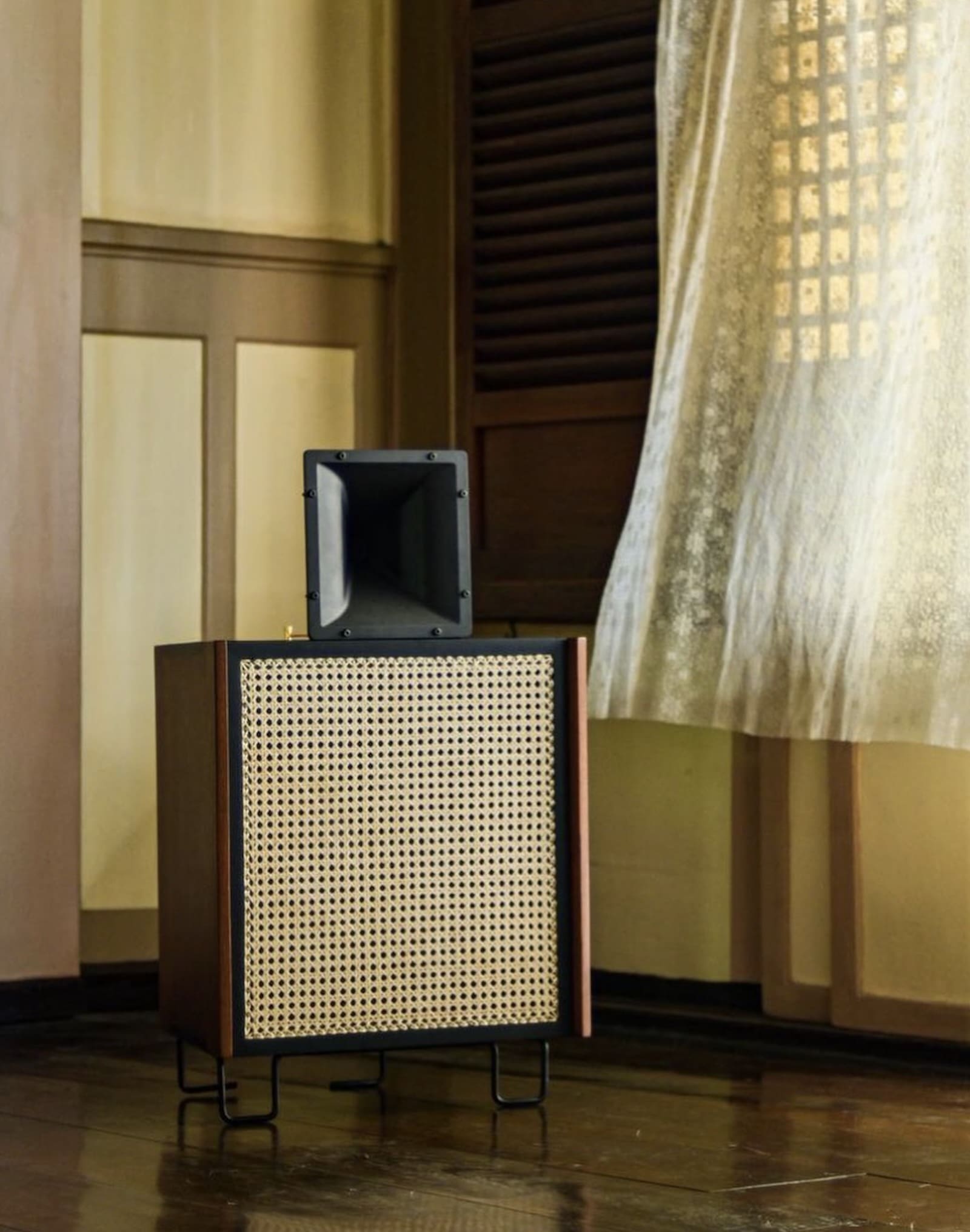 A mid-size brown and beige sound speaker standing in the corner of a room, next to a window with a curtain that has caught a slight breeze