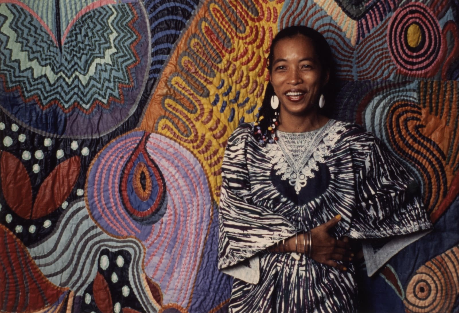 Artist Pacita Abad, wearing a black and white dress, stands in front of one of her wall-sized, colorful paintings