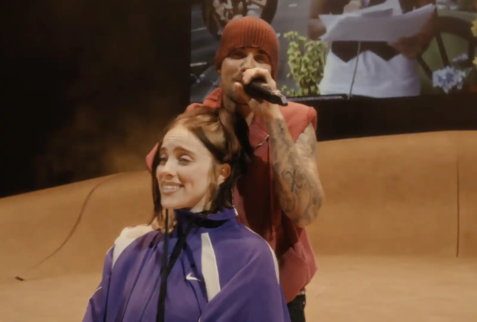 Male performer in a red beanie holds a microphone behind a smiling woman seated in a purple cape at a live stage event, as if giving a haircut.