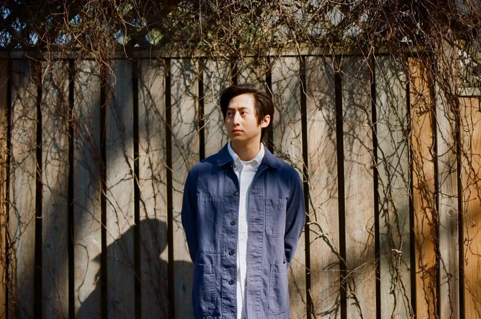 Young man in a blue jacket and white shirt stands in front of a vine-covered fence, looking to the right.