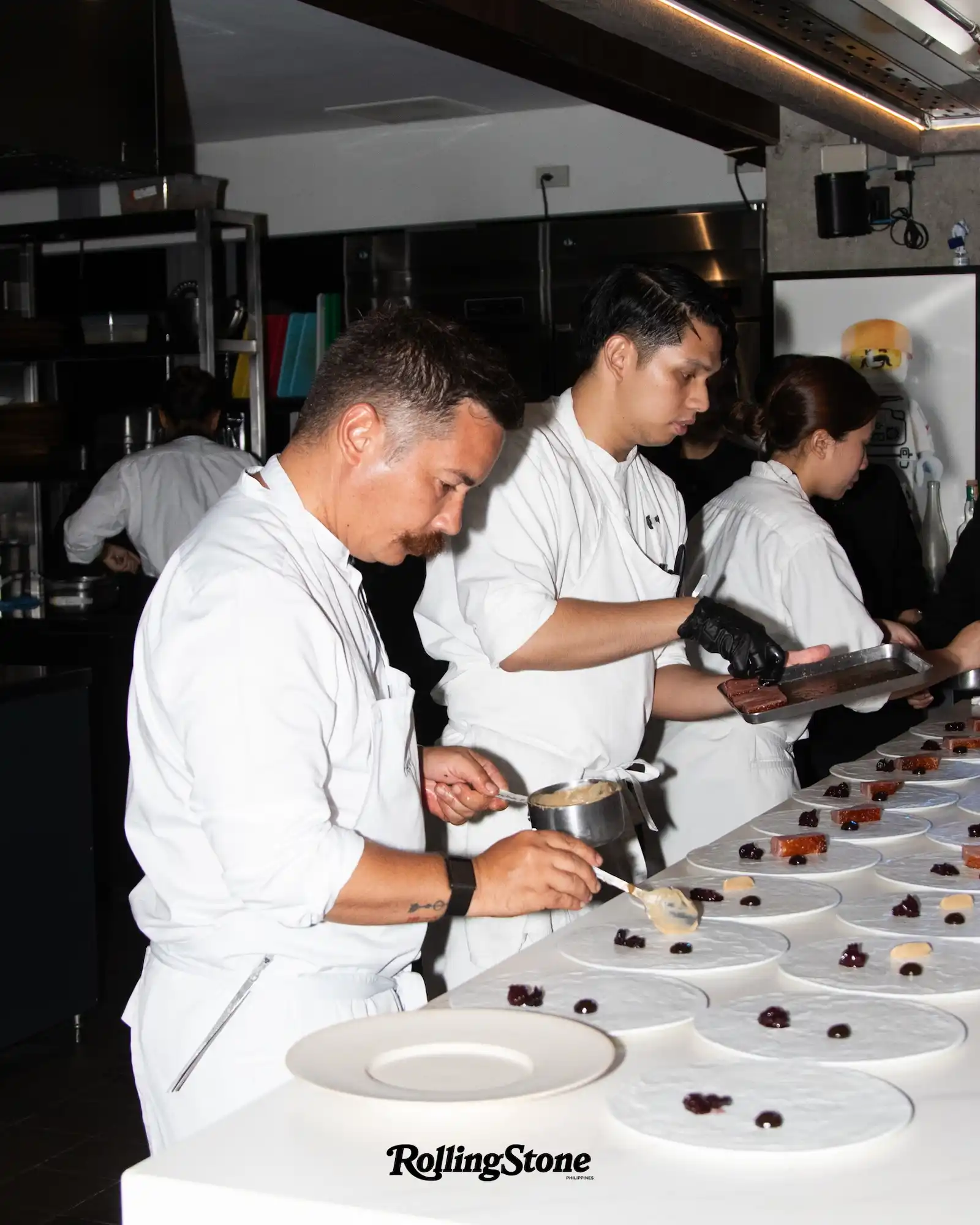 Chefs in white uniforms plate small desserts along a long row of plates in a kitchen; Rolling Stone logo visible in foreground.