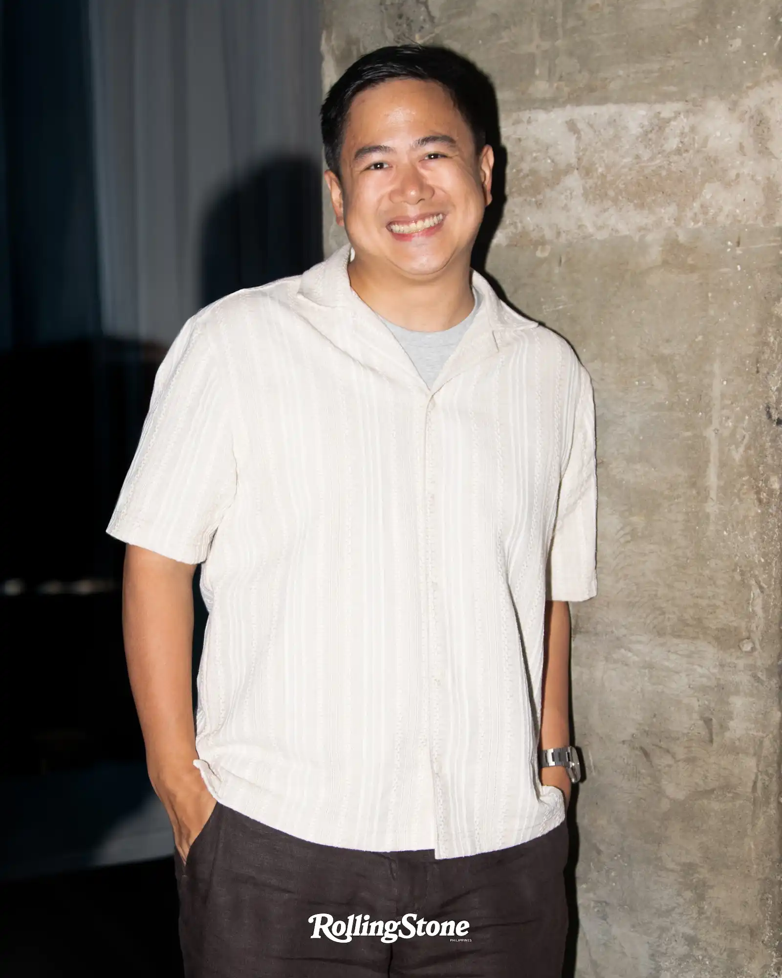 Smiling man in a pale striped shirt with hands in pockets, standing beside a rough concrete wall; Rolling Stone logo at the bottom.