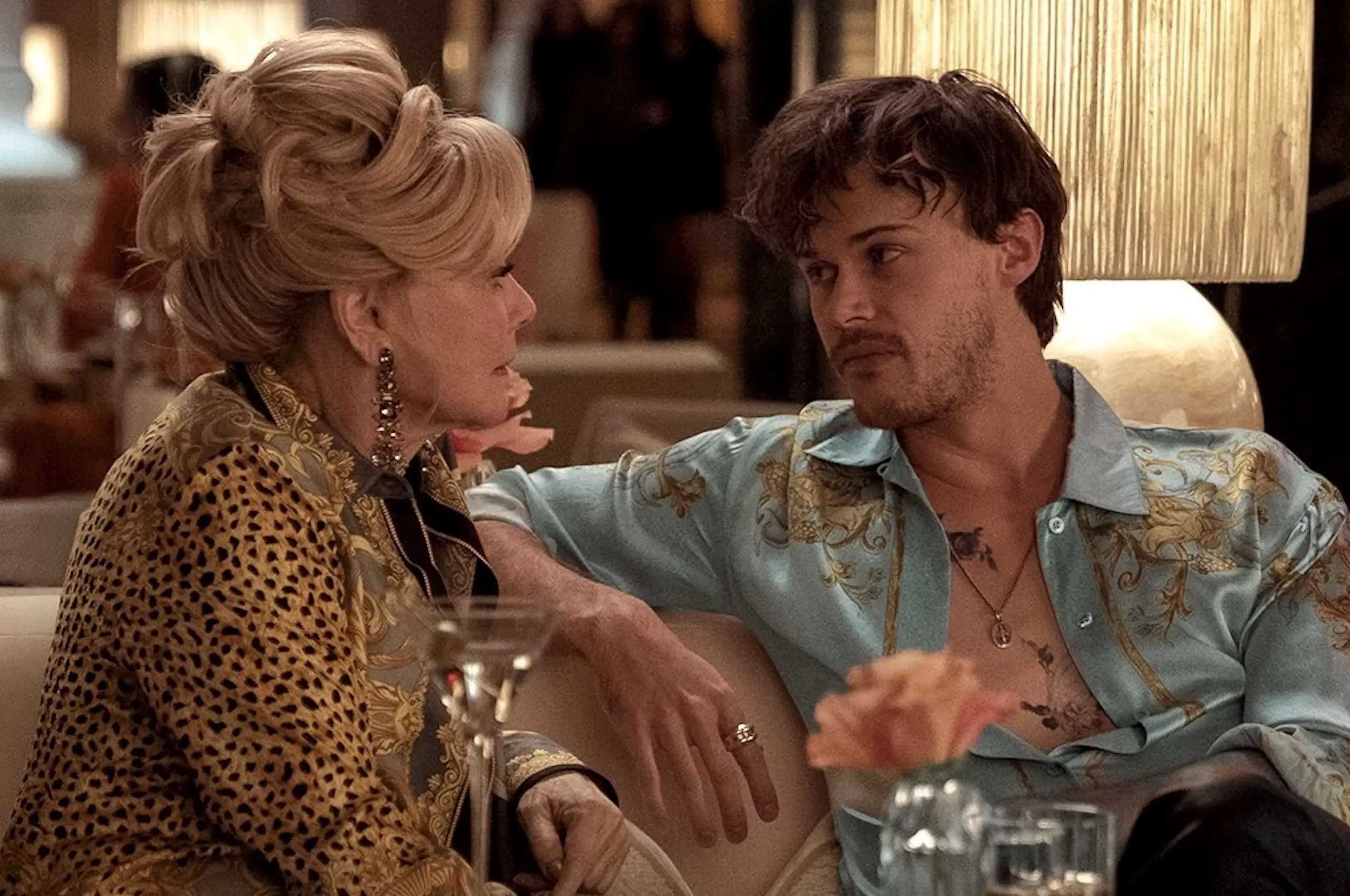 Older woman in a leopard-print outfit talking with a younger man in a tropical shirt at a dinner table, leaning in toward each other.