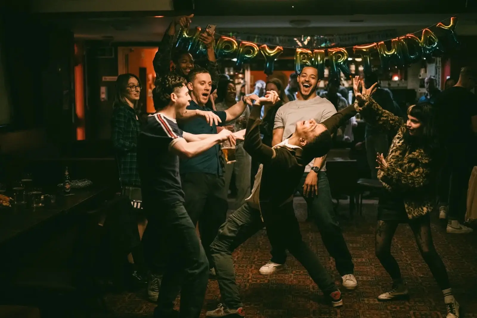 Group of friends dancing and laughing at a birthday party under a 'Happy Birthday' banner.