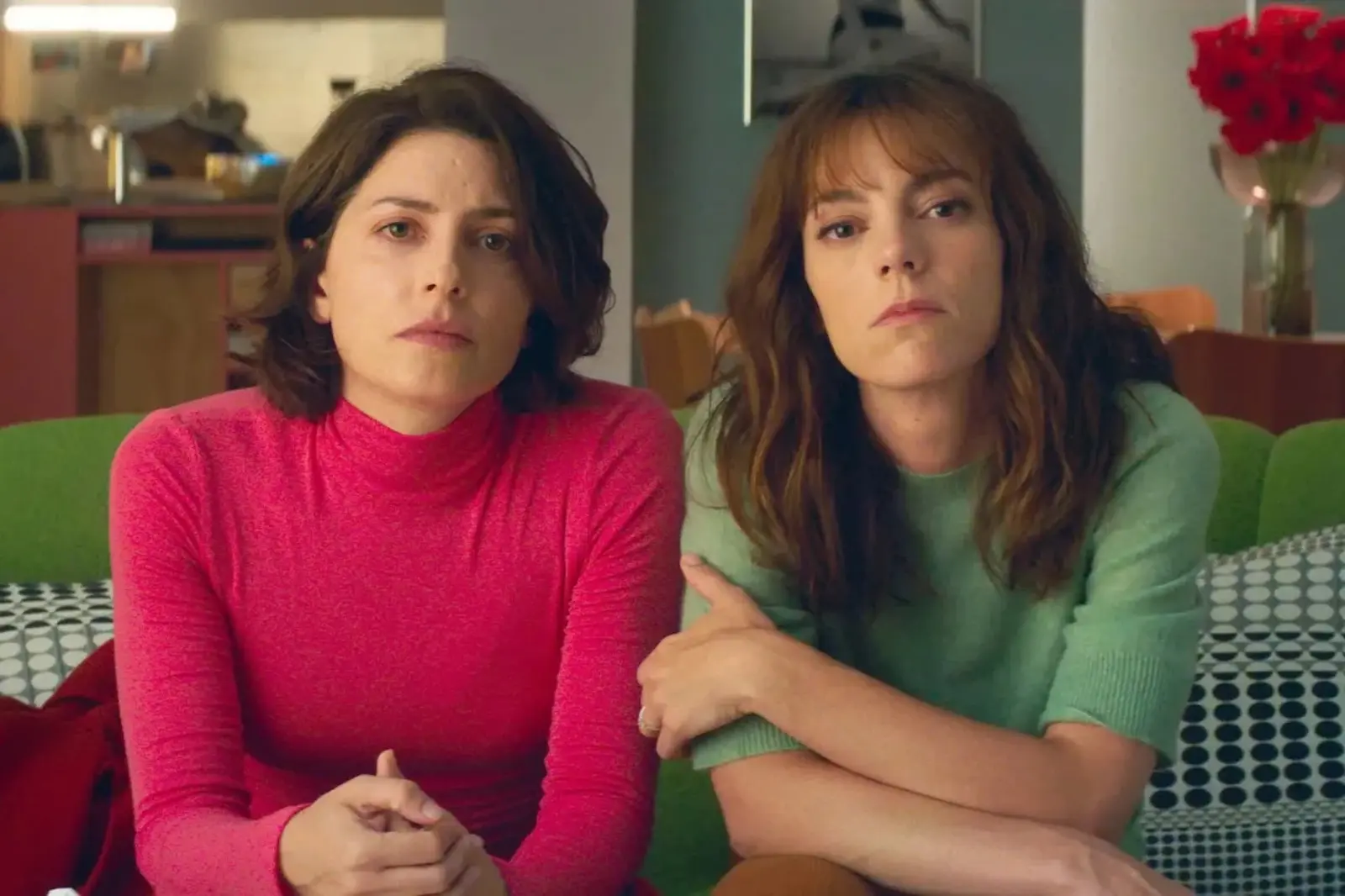 Two women sit closely on a green sofa, looking off to the side; one wears a red turtleneck, the other a light green sweater in a cozy living room.
