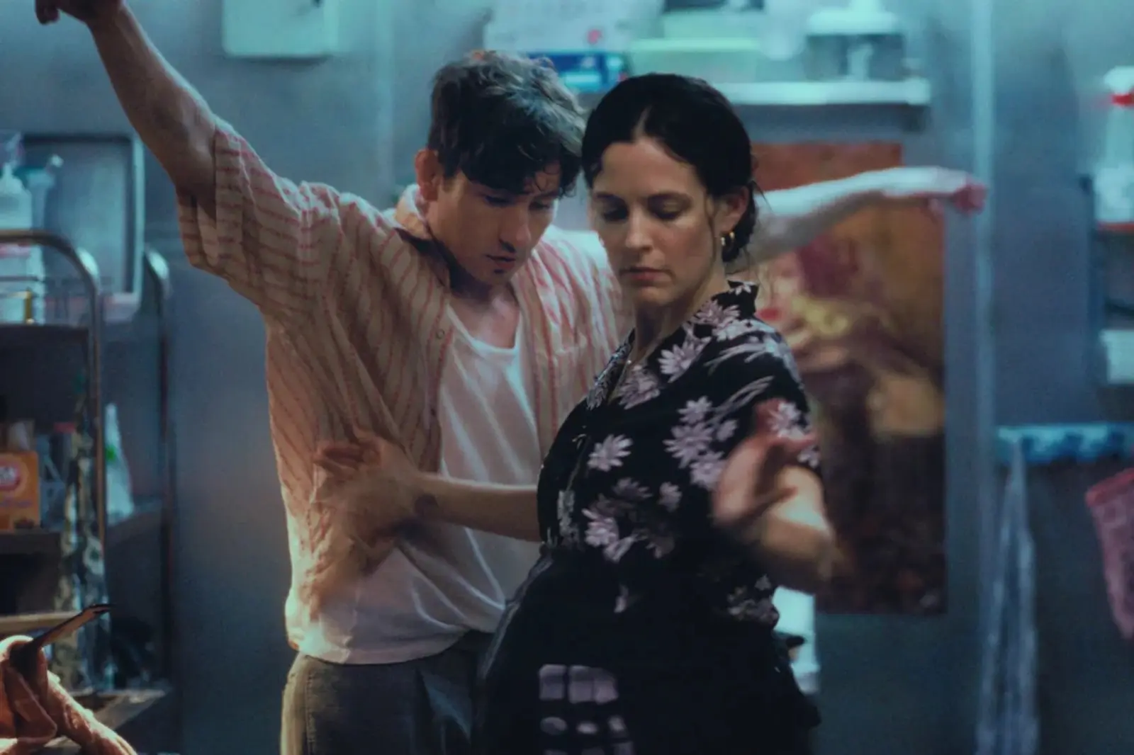 A man and woman dance closely in a dimly lit kitchen, arms raised and focused expressions.