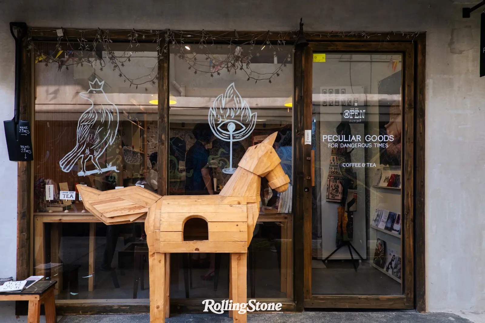 Wooden horse sculpture in front of a glass storefront with illustrated logos on the glass and a 'PECULIAR GOODS FOR DANGEROUS TIMES COFFEE & TEA' sign inside; Rolling Stone logo at bottom edge of the window.