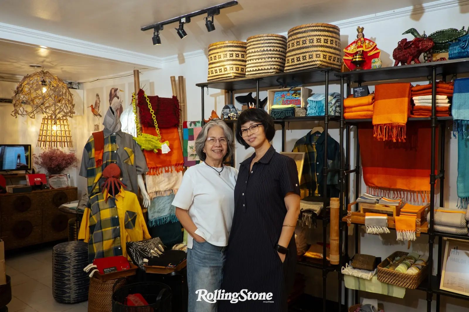 Two smiling women pose together in a colorful textile shop, surrounded by woven baskets, blankets, scarves, and clothing on shelves.