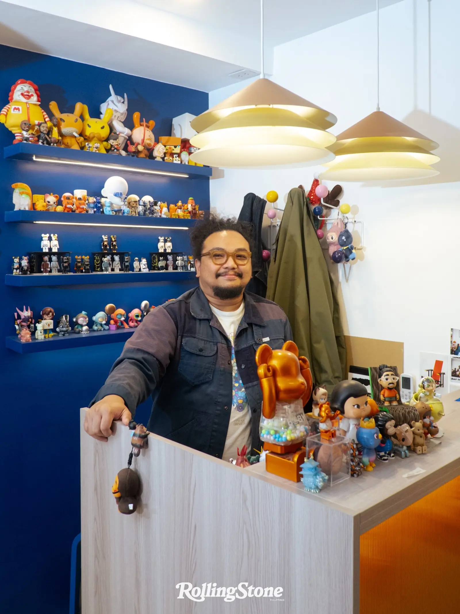 A man wearing glasses smiles behind a toy display counter filled with colorful figurines in a blue-walled shop.