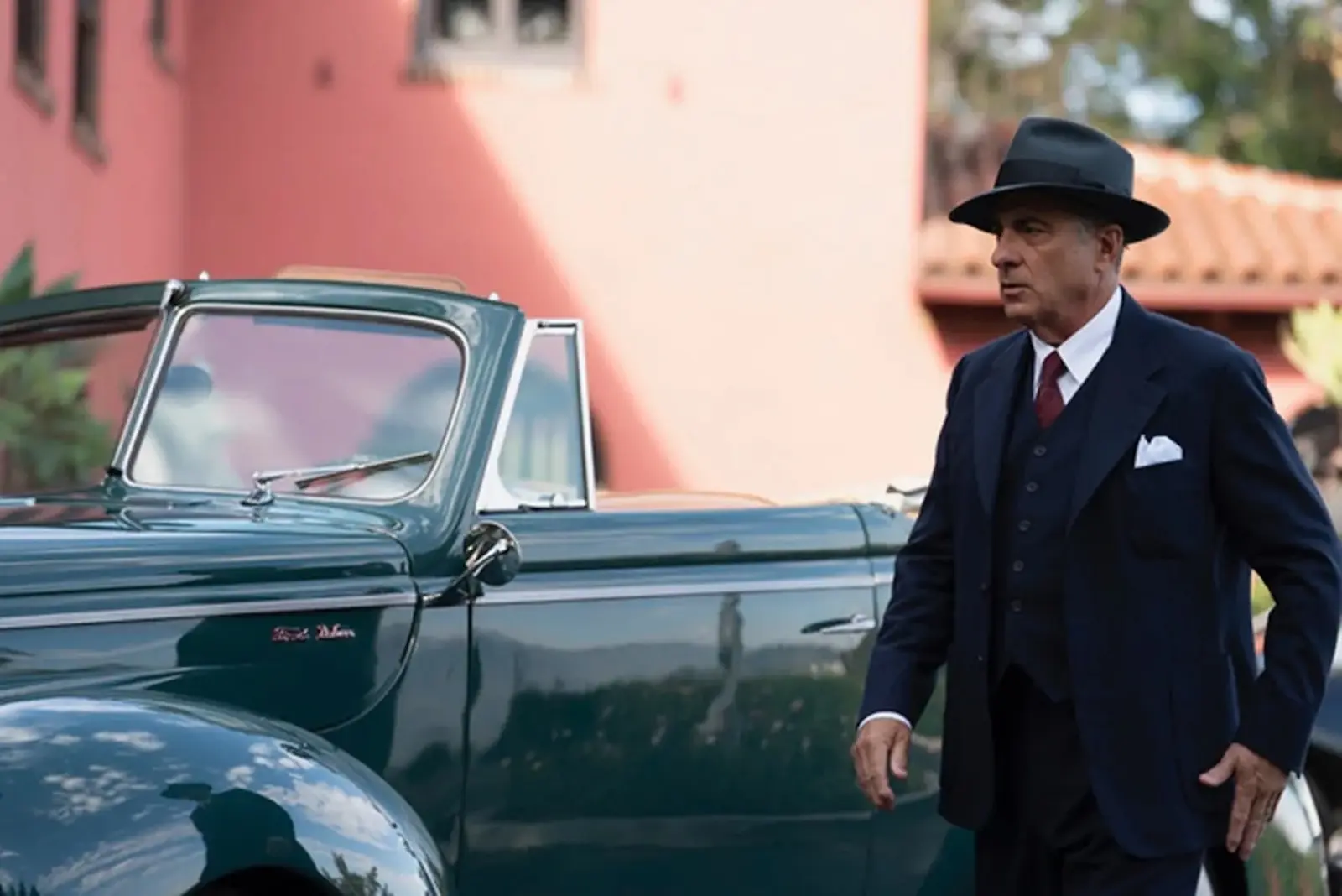 Man in a dark suit and fedora stands beside a green vintage convertible car.
