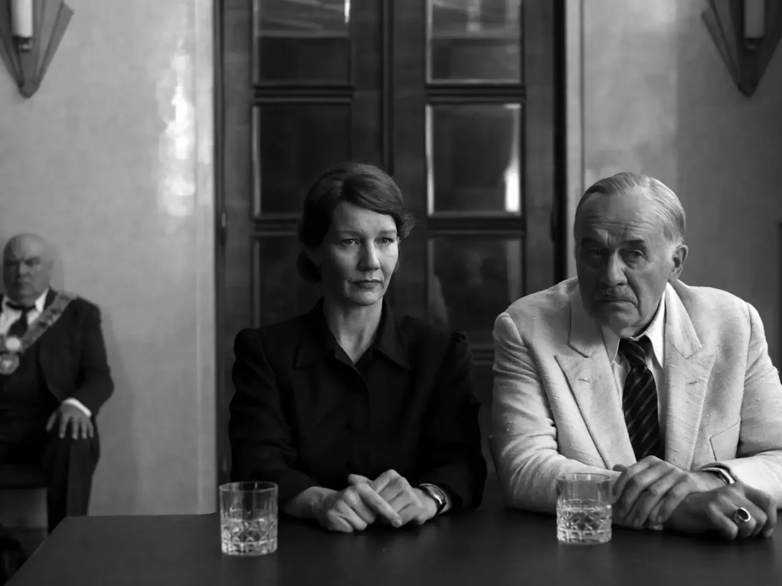Two serious-looking people sit at a table with glasses in front of them; a woman in a dark blazer and a man in a light suit lean forward, while a third person sits blurred in the background.