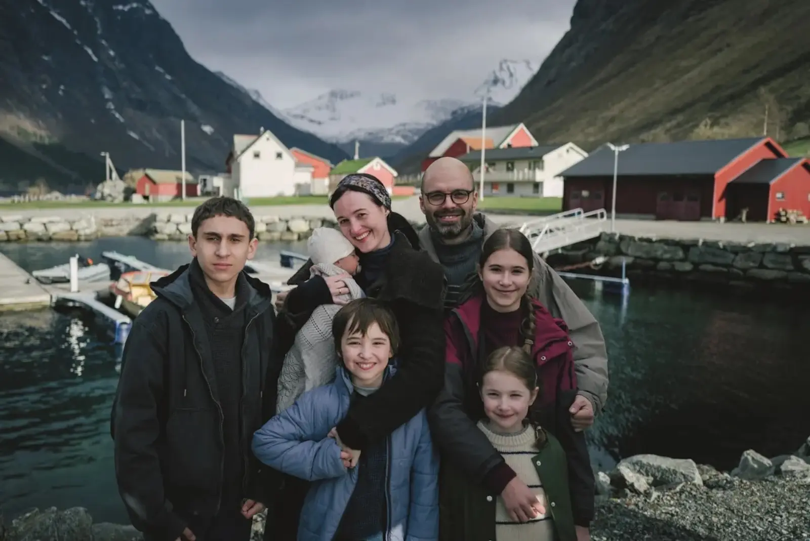 A family of seven posing together by a small harbor with colorful houses and mountains in the background.
