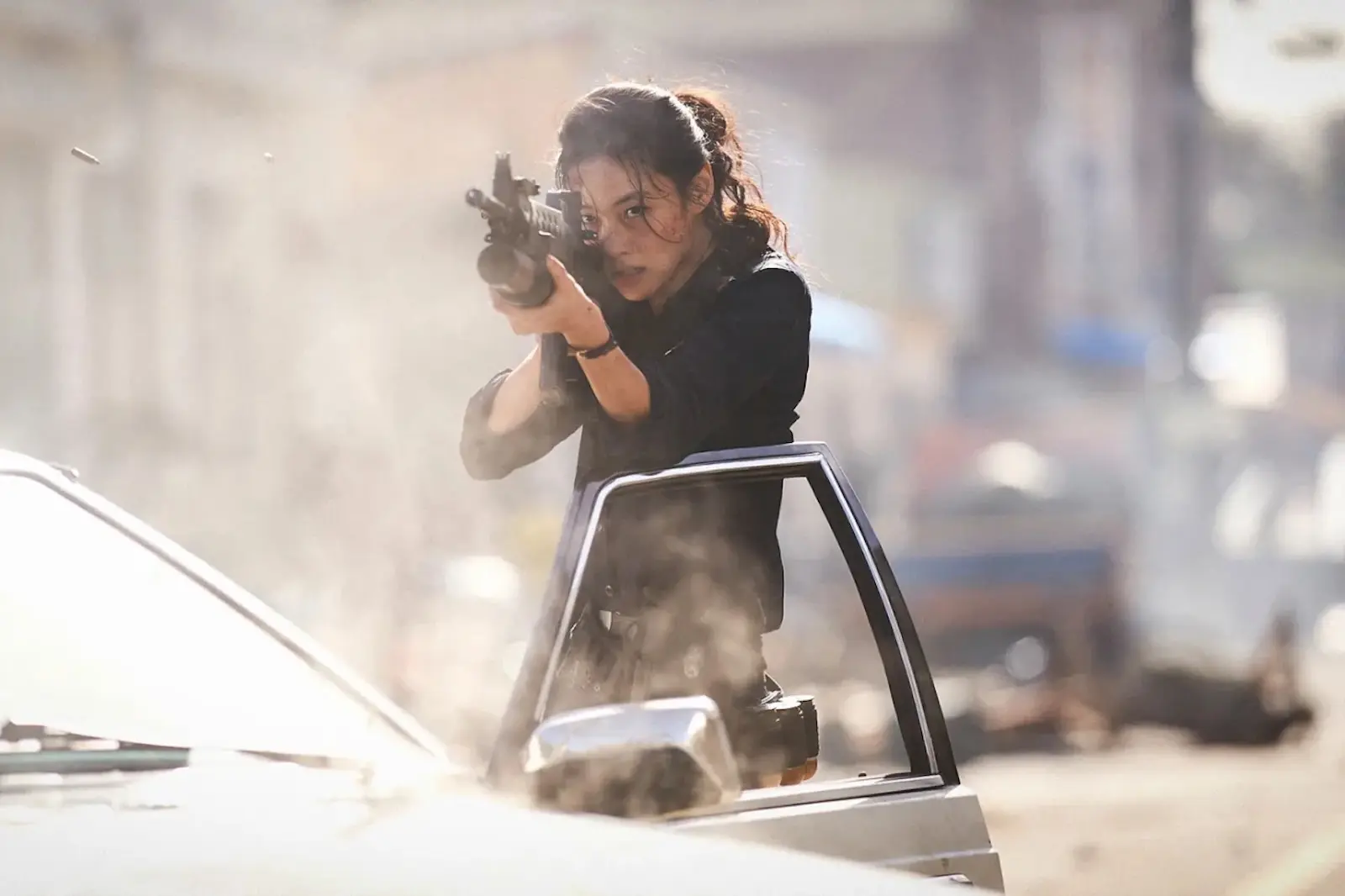 Woman aiming a rifle from a car window during a dusty street action scene.