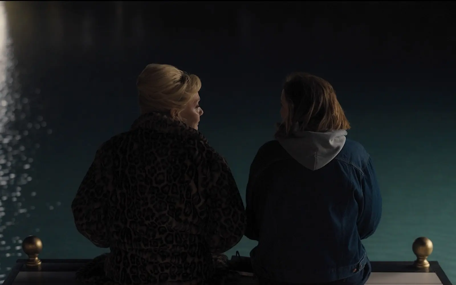 Two people in warm jackets sit on a railing overlooking a dark, moonlit body of water at night Hacks S05E06 deborah ava fur coat