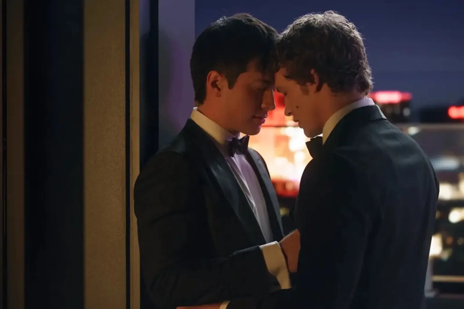 Two men in tuxedos share an intimate moment, foreheads touching, against a backdrop of city lights at night.