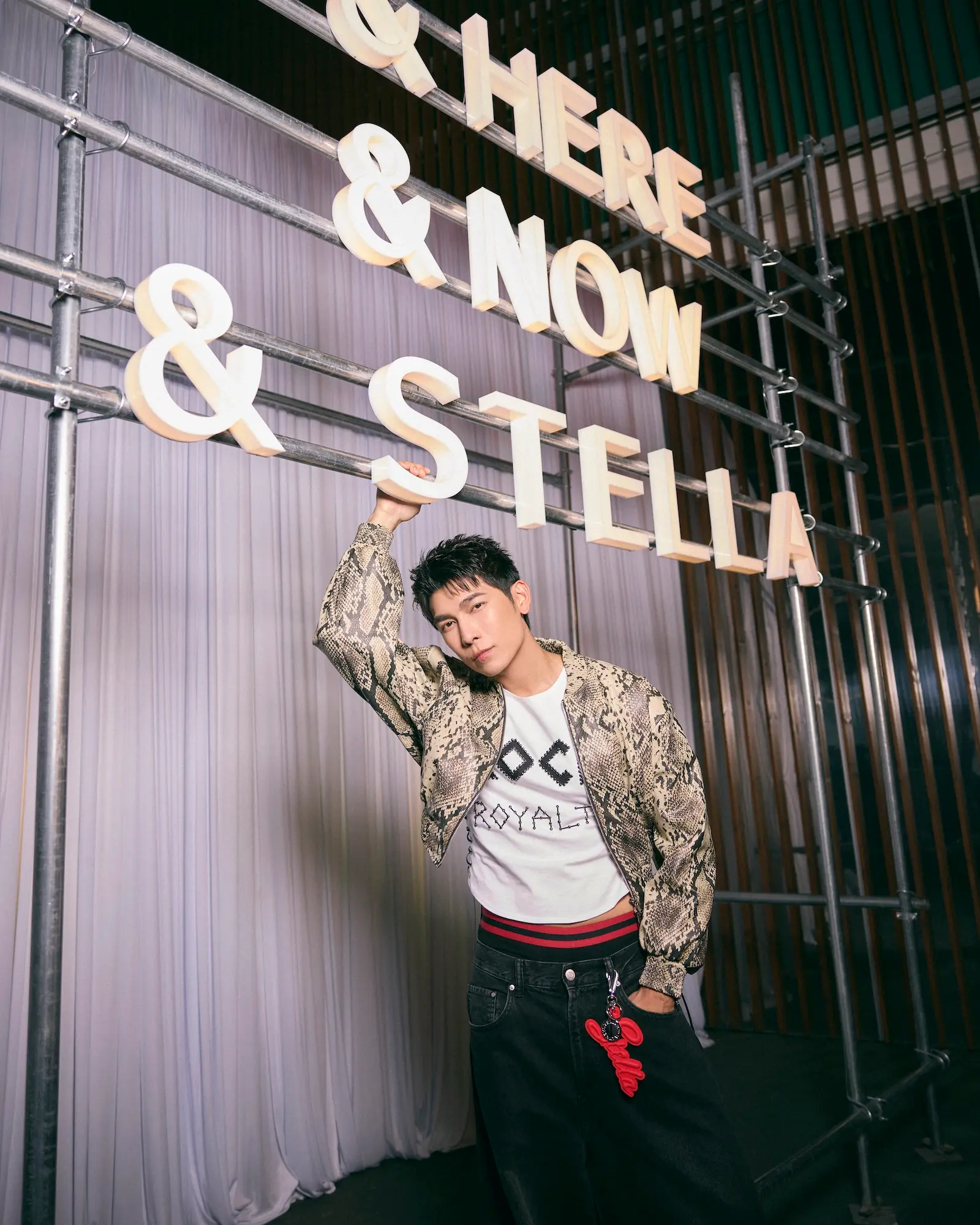 Young man in a snakeskin jacket poses under giant white letters on a metal scaffolding backdrop in a studio setting.