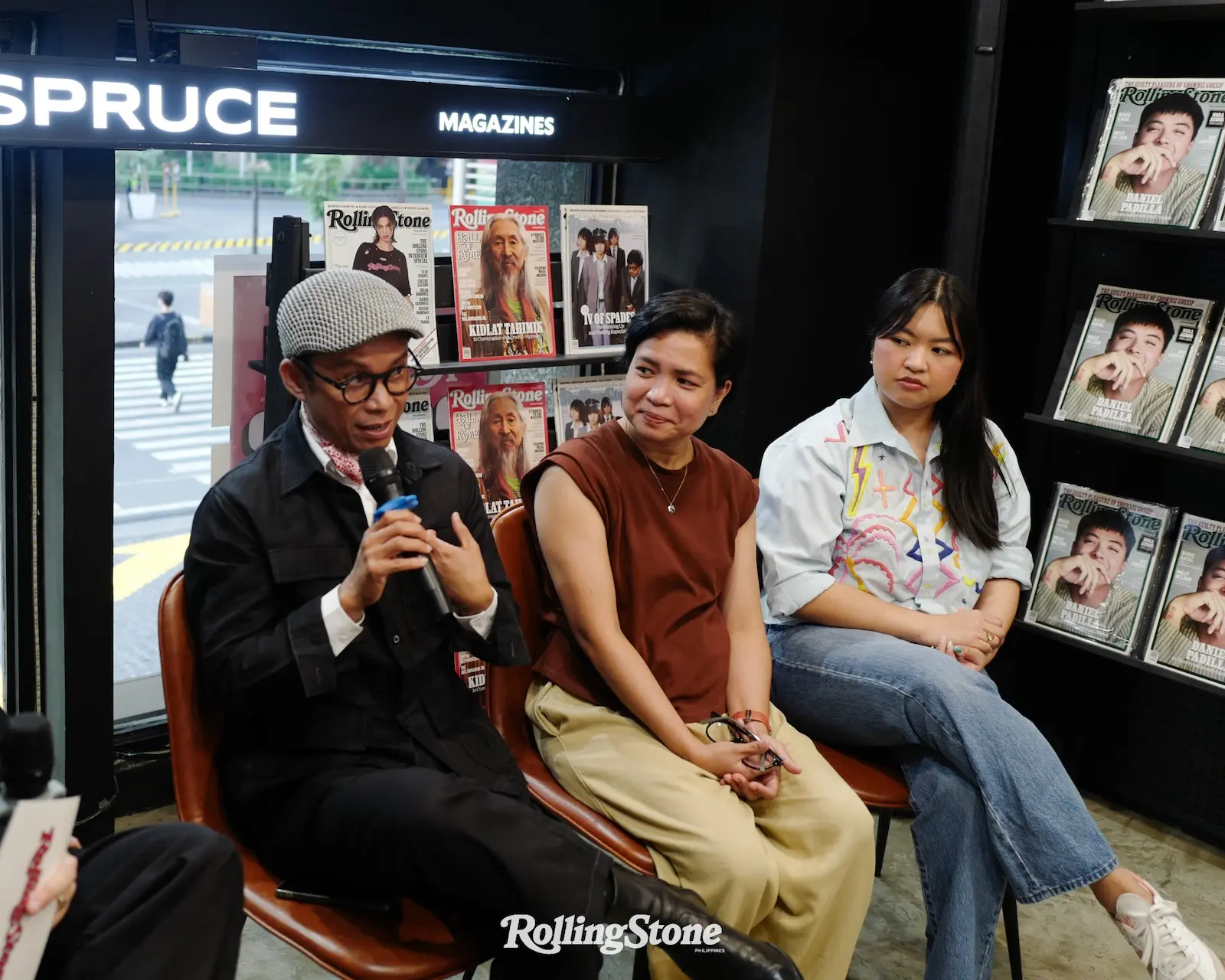 Three people sit in a magazine shop during a panel discussion, Rolling Stone magazines on display behind them.