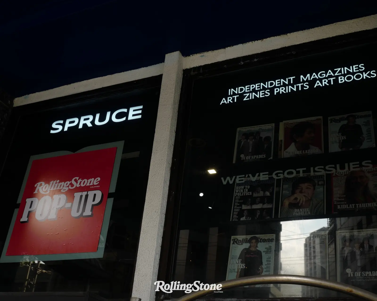 Storefront window for a magazine shop with a bright 'SPRUCE' sign and a red Rolling Stone poster inside/nearby.