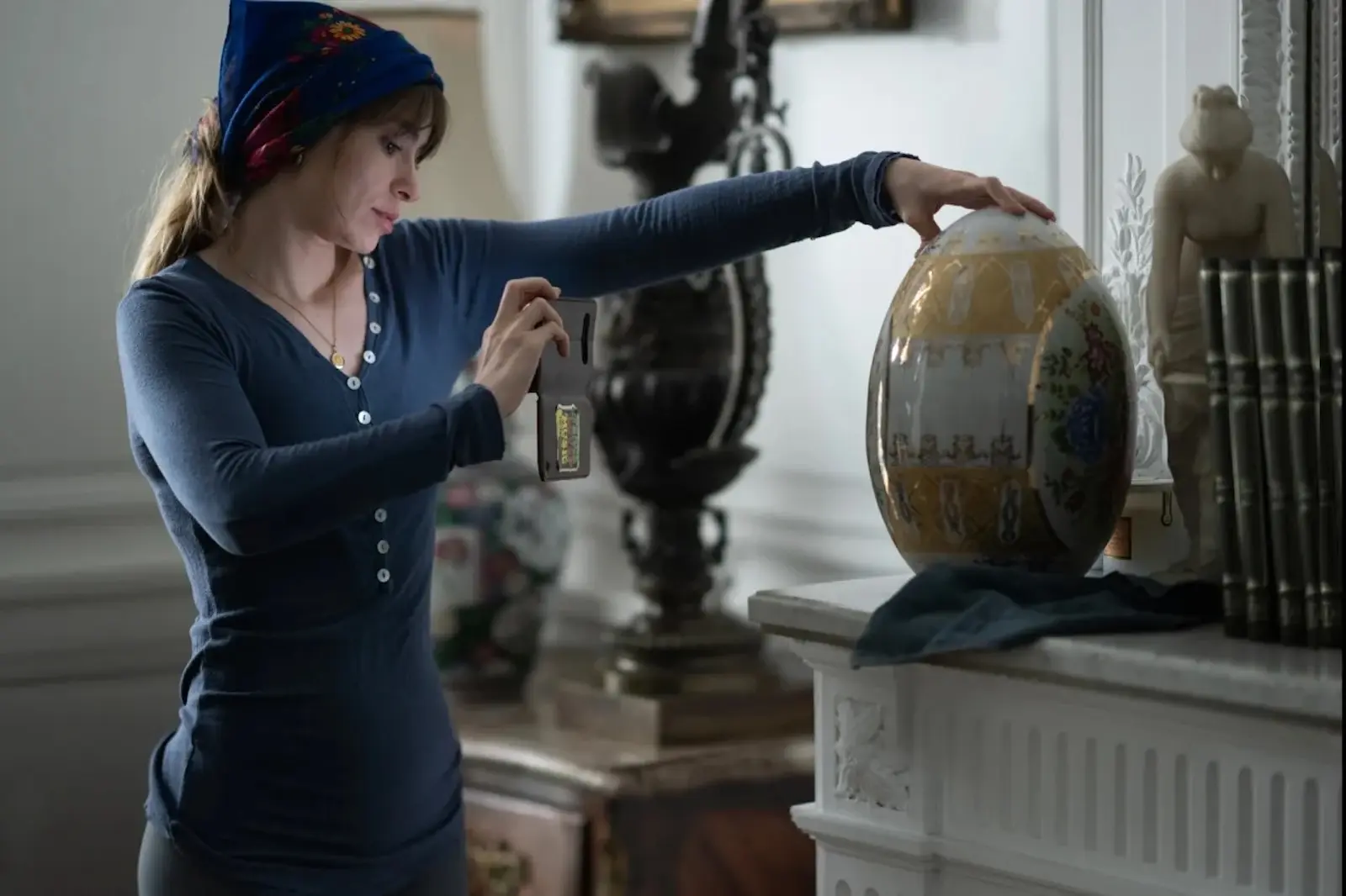 Woman in a blue long-sleeve shirt and blue headscarf attaches a tag to a large decorated egg sculpture on a mantel.