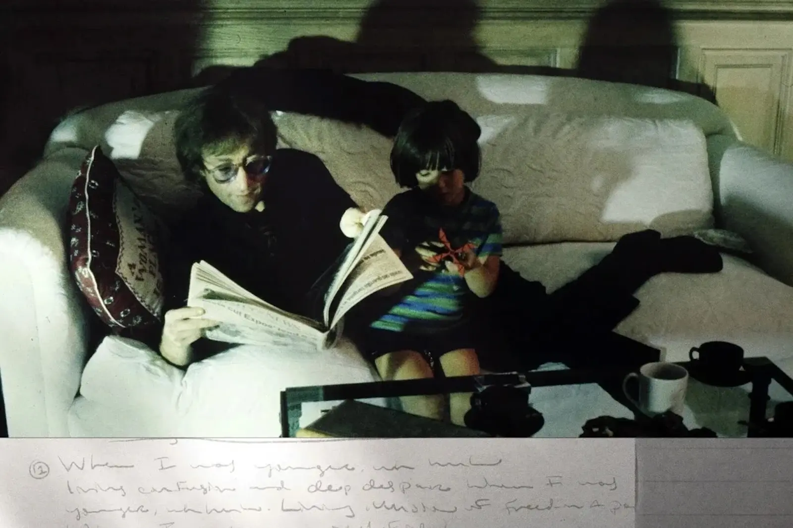 Two people sit on a white couch: an adult reading a newspaper, a child beside them looking at a device or paper in a dim living room setting with a coffee table in front.