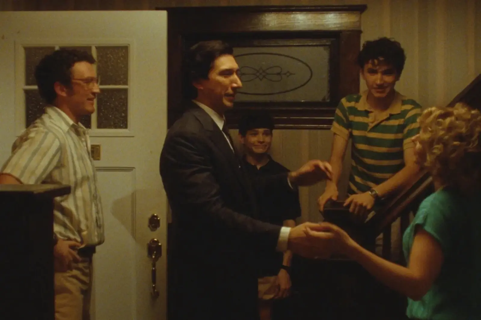 Group of adults inside a home greeting each other; a man in a suit shakes hands with a woman on the right as others watch.