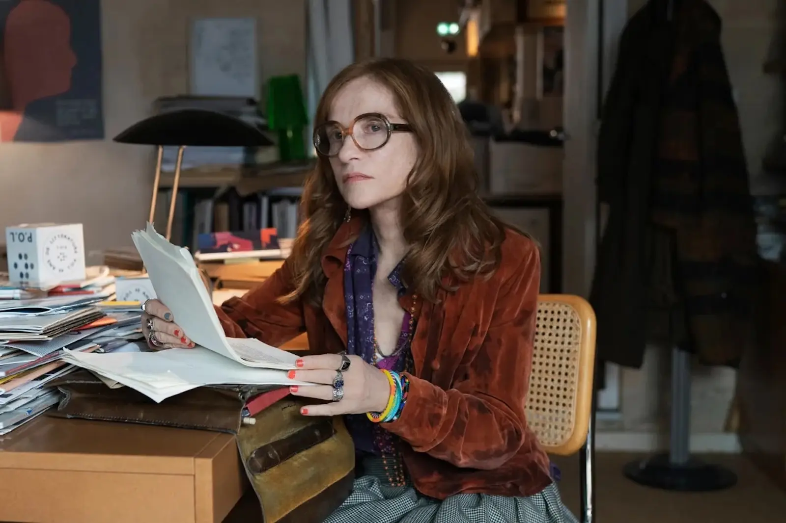 Woman with round glasses and brown hair reads a stack of papers at a cluttered desk.
