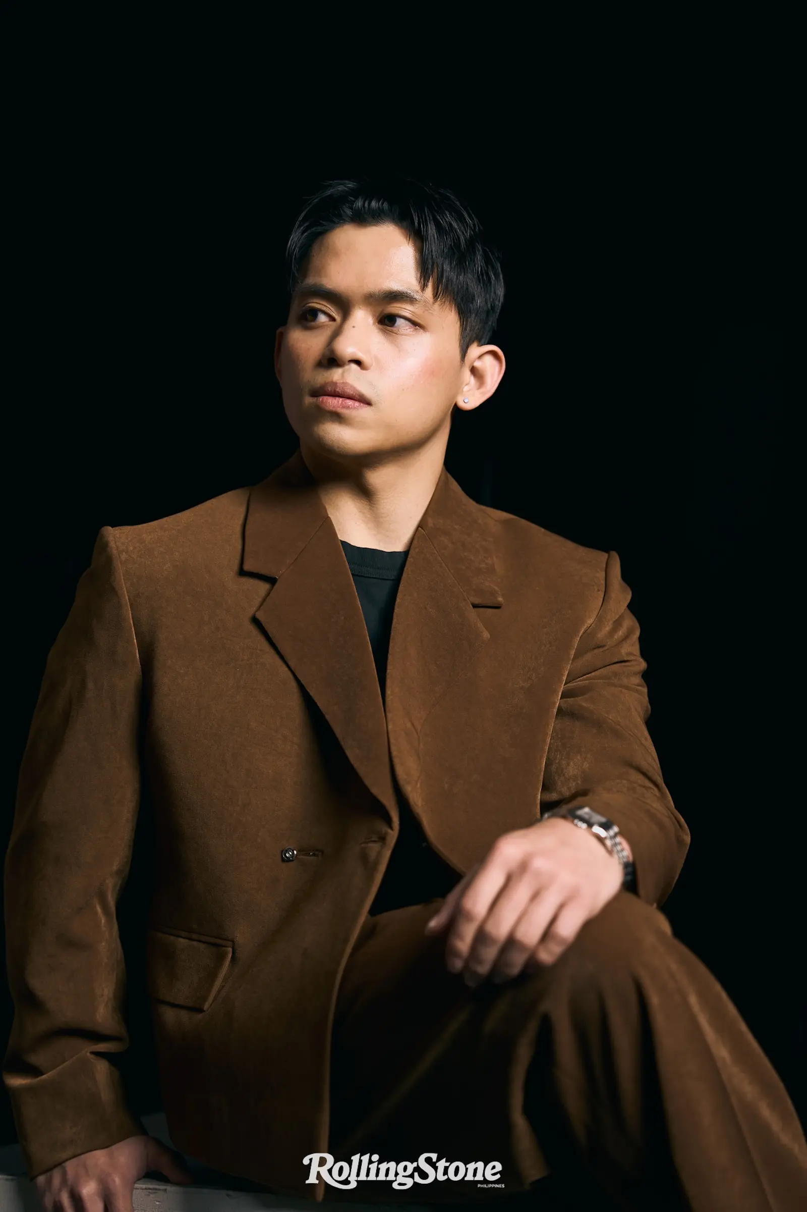 Portrait of a man wearing a brown blazer, looking to the left against a dark background.