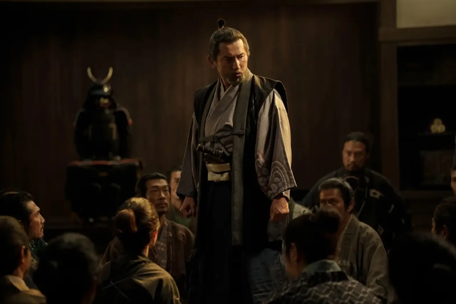 Man in traditional kimono stands on a platform addressing a crowd of seated samurai.