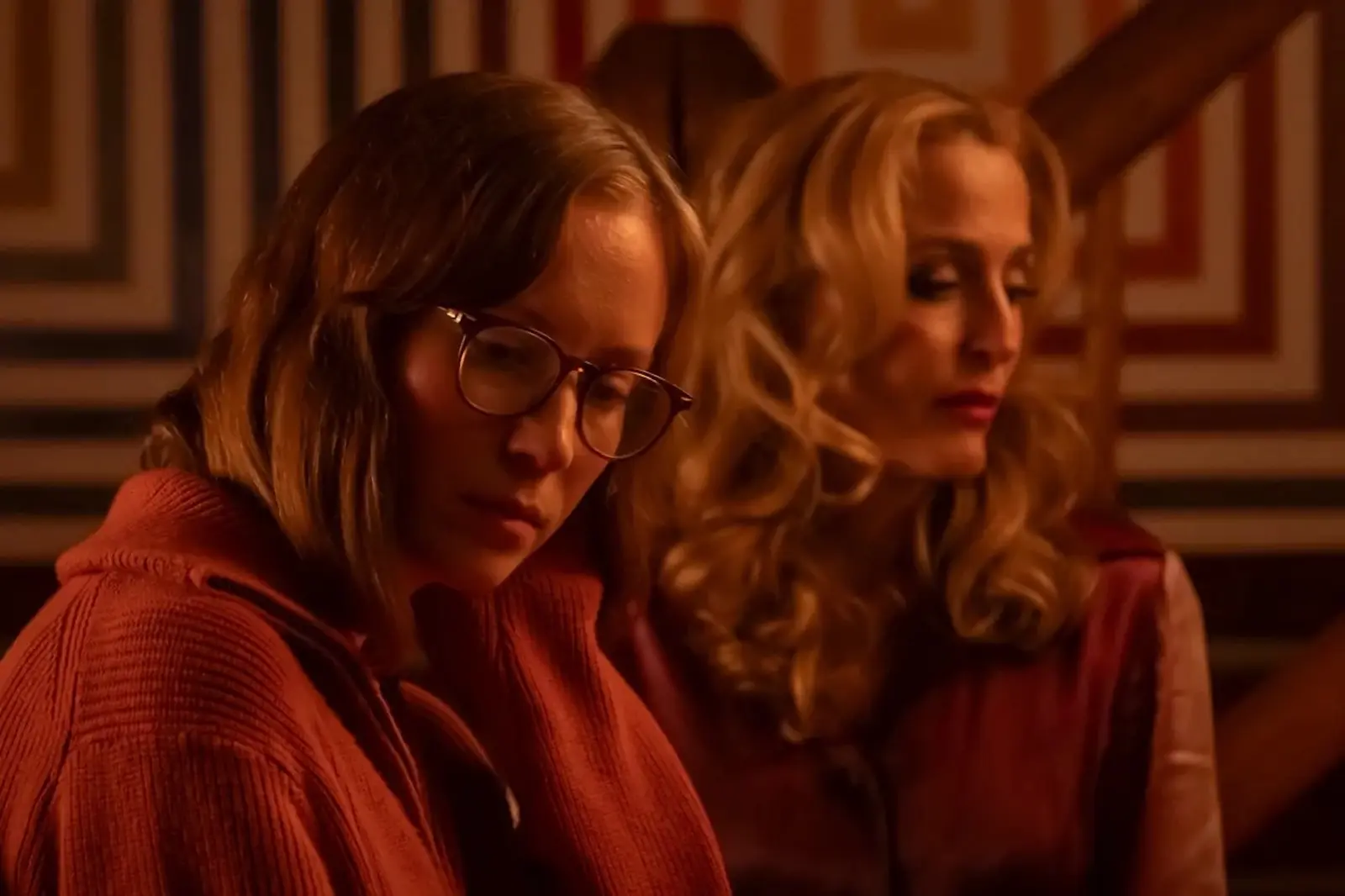 Two women sit indoors in warm, amber lighting; the woman on the left wears glasses and looks down, while the woman on the right looks to the side with wavy blonde hair.