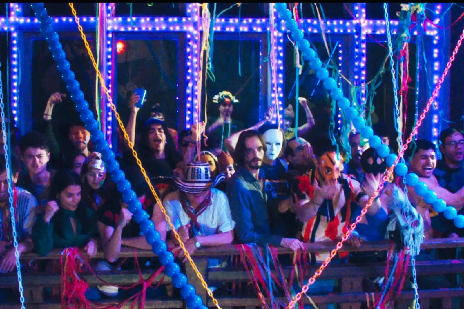 Crowd at a neon-lit party dancing behind a barrier with colorful streamers and blue LED lights gradually illuminating the scene.