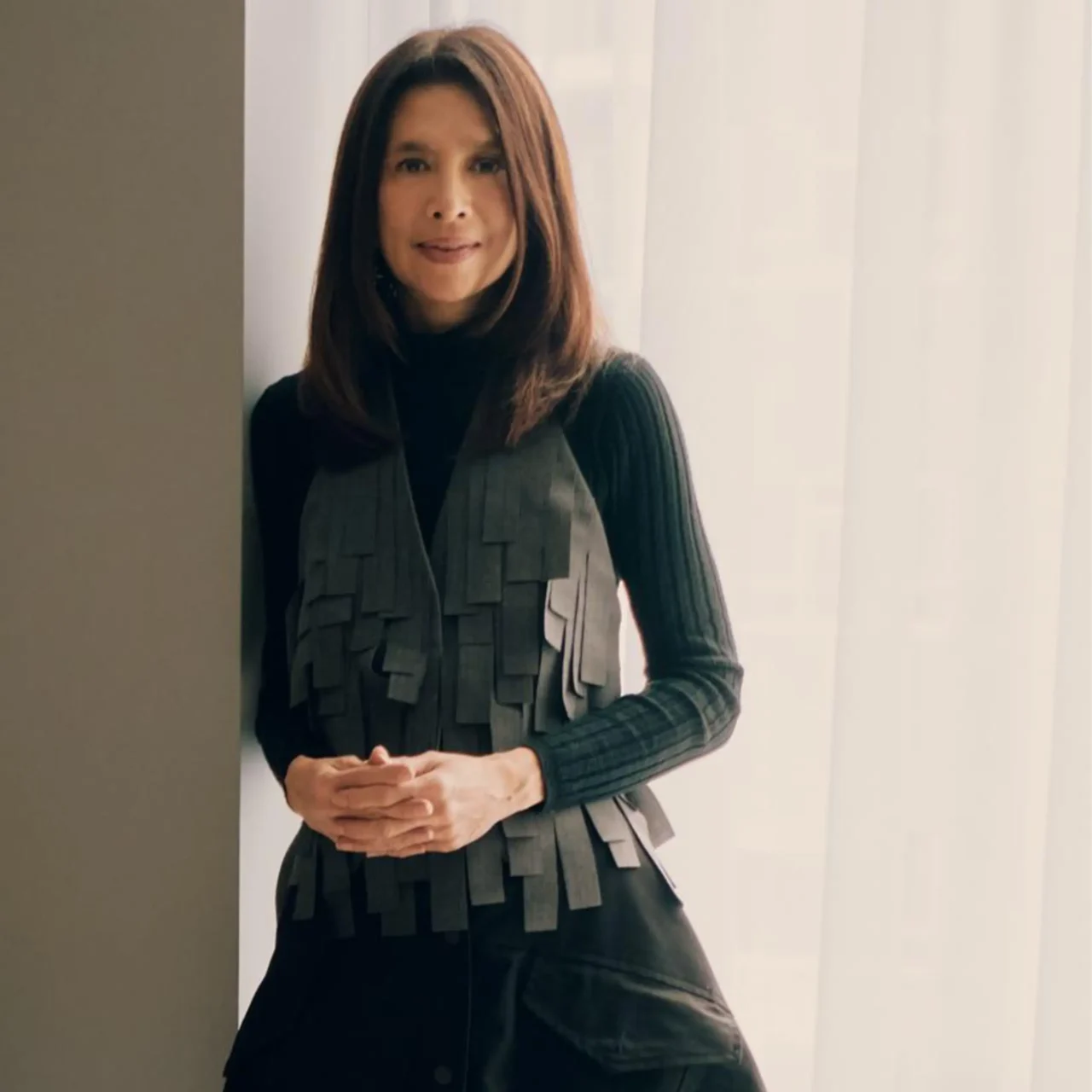 Portrait of a woman with shoulder-length brown hair, wearing a black turtleneck and a gray layered vest, standing indoors by a doorway/curtain.”,