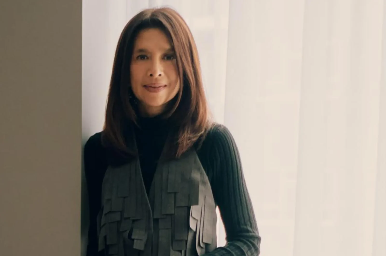 Portrait of a woman with shoulder-length brown hair, wearing a black turtleneck and a gray layered vest, standing indoors by a doorway/curtain.”,