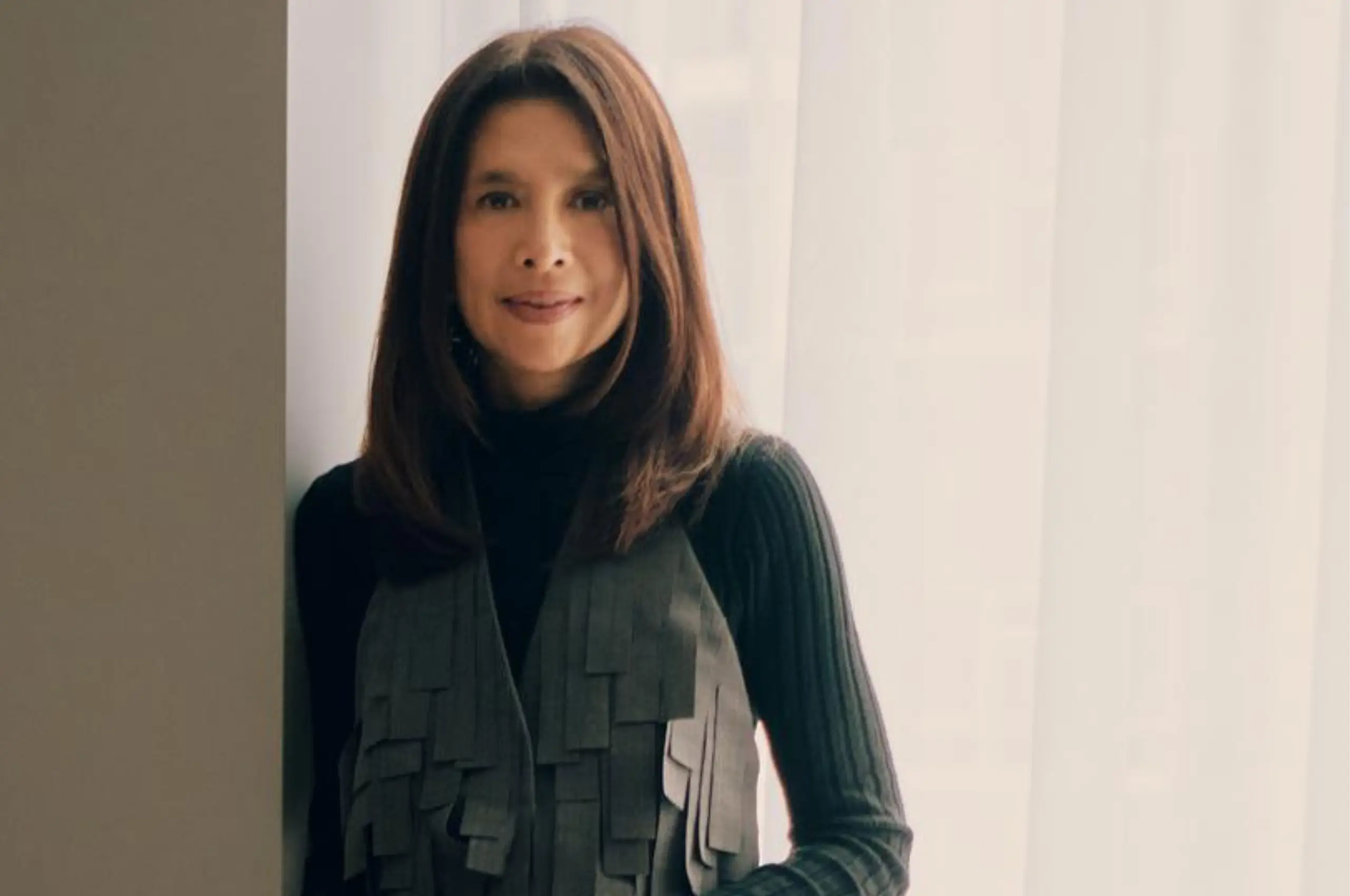 Portrait of a woman with shoulder-length brown hair, wearing a black turtleneck and a gray layered vest, standing indoors by a doorway/curtain.”,