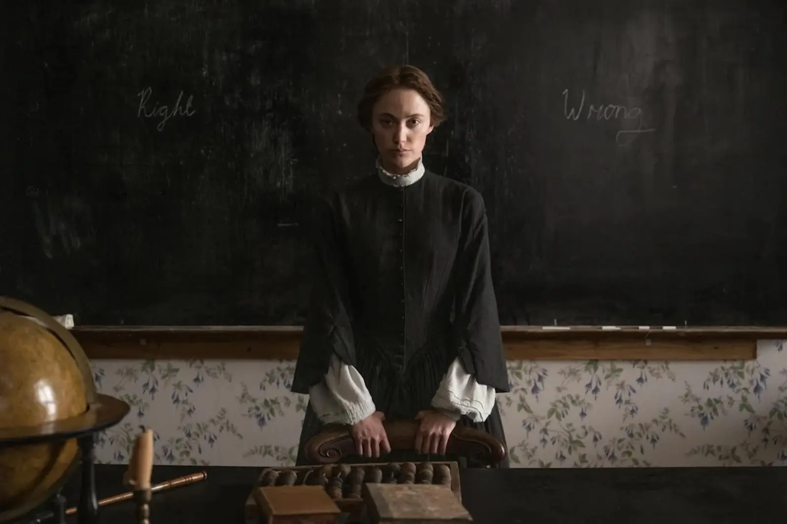 Woman in a dark period dress standing at a classroom desk, with a globe and chalkboard behind her.