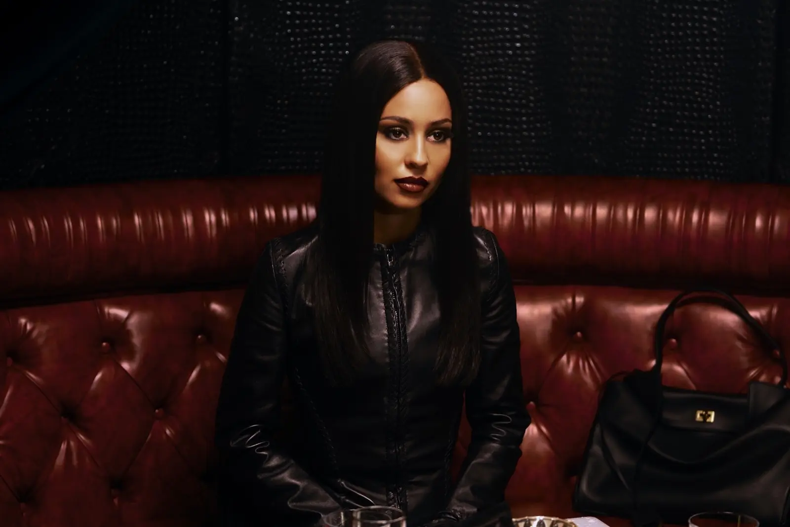 Woman with long dark hair in a black leather jacket seated on a red tufted booth in a dim lounge, looking forward with a neutral expression, handbag beside her.