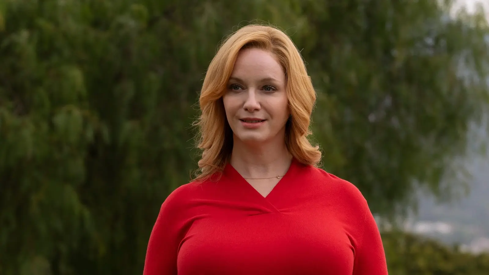 Woman with shoulder-length red hair in a red top standing outdoors with greenery behind her. hacks christina hendricks