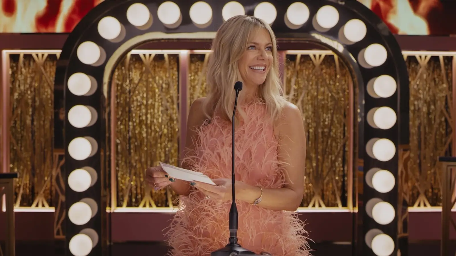 Woman in a pink feathered dress hosting at a glamorous stage, standing behind a microphone and smiling. hacks kaitlin olson dj vance