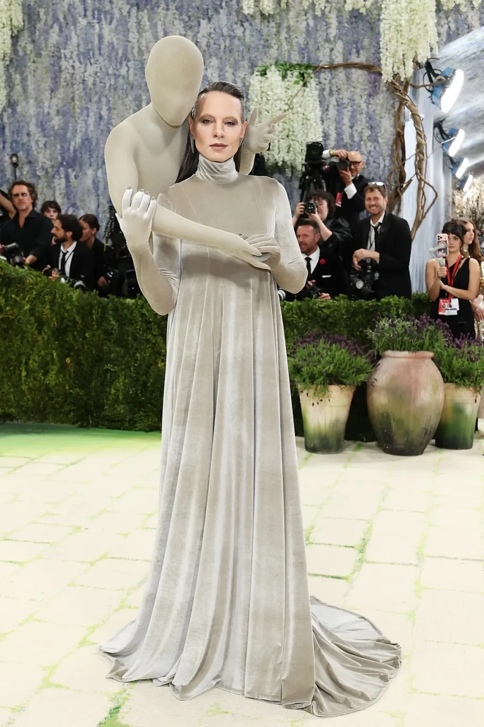 A person in a floor-length silver-gray gown poses with a large, featureless statue behind them at a fashion event with photographers in the background.