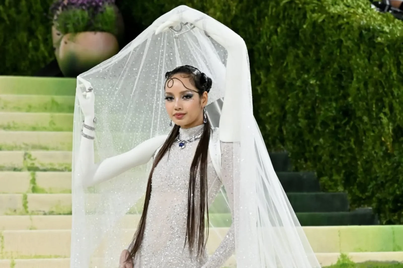 Woman in a glittering silver gown and long white veil posing on outdoor stone steps with greenery behind her.
