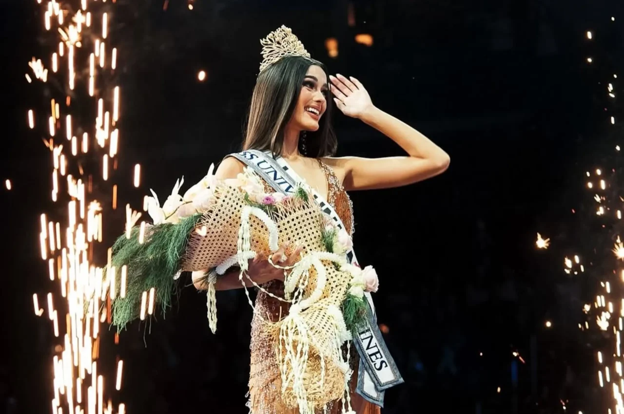 Miss pageant winner on stage, wearing crown and sash, holding a bouquet as gold sparks rise around her.