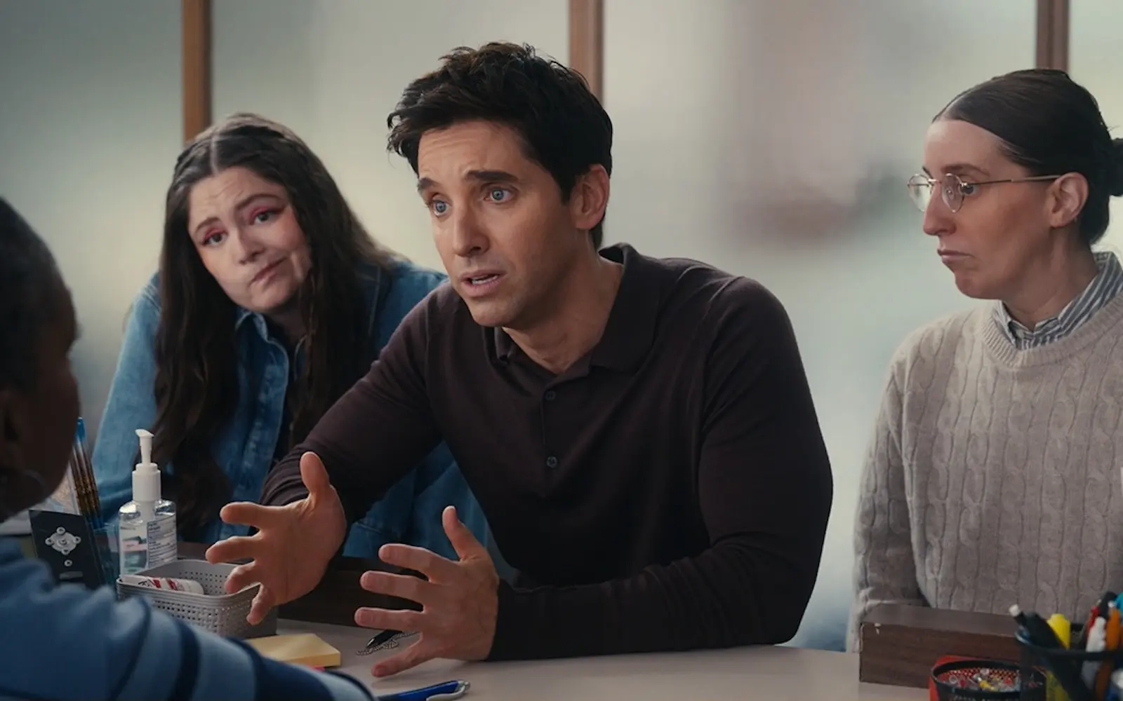 paul w downs meg stalter robbie hoffman hacks season 5 episode 9 Man in a dark polo explains something at a table, gesturing with hands, flanked by two attentive women in a meeting setting.