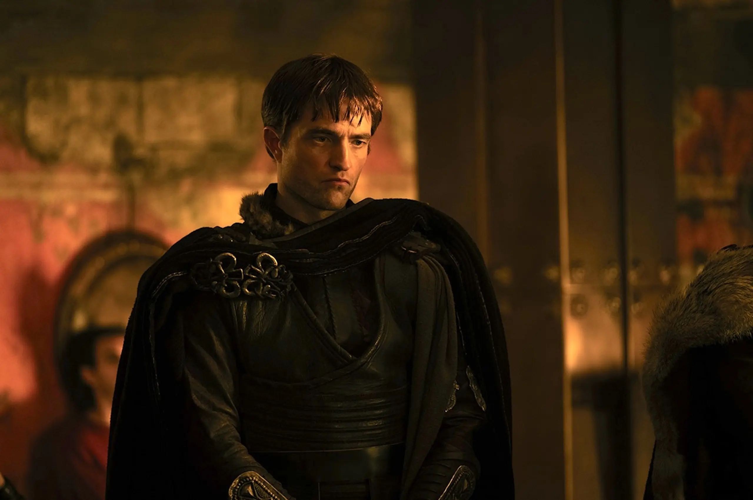 Man in a dark medieval cloak with fur trim stands in a warmly lit, medieval interior, looking contemplative to the side.