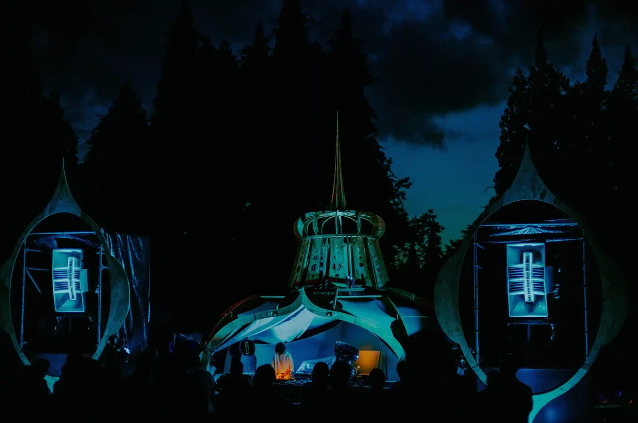 Nighttime outdoor art installation with glowing blue structures and a crowd silhouetted in front of dark trees and tents at center stage.
