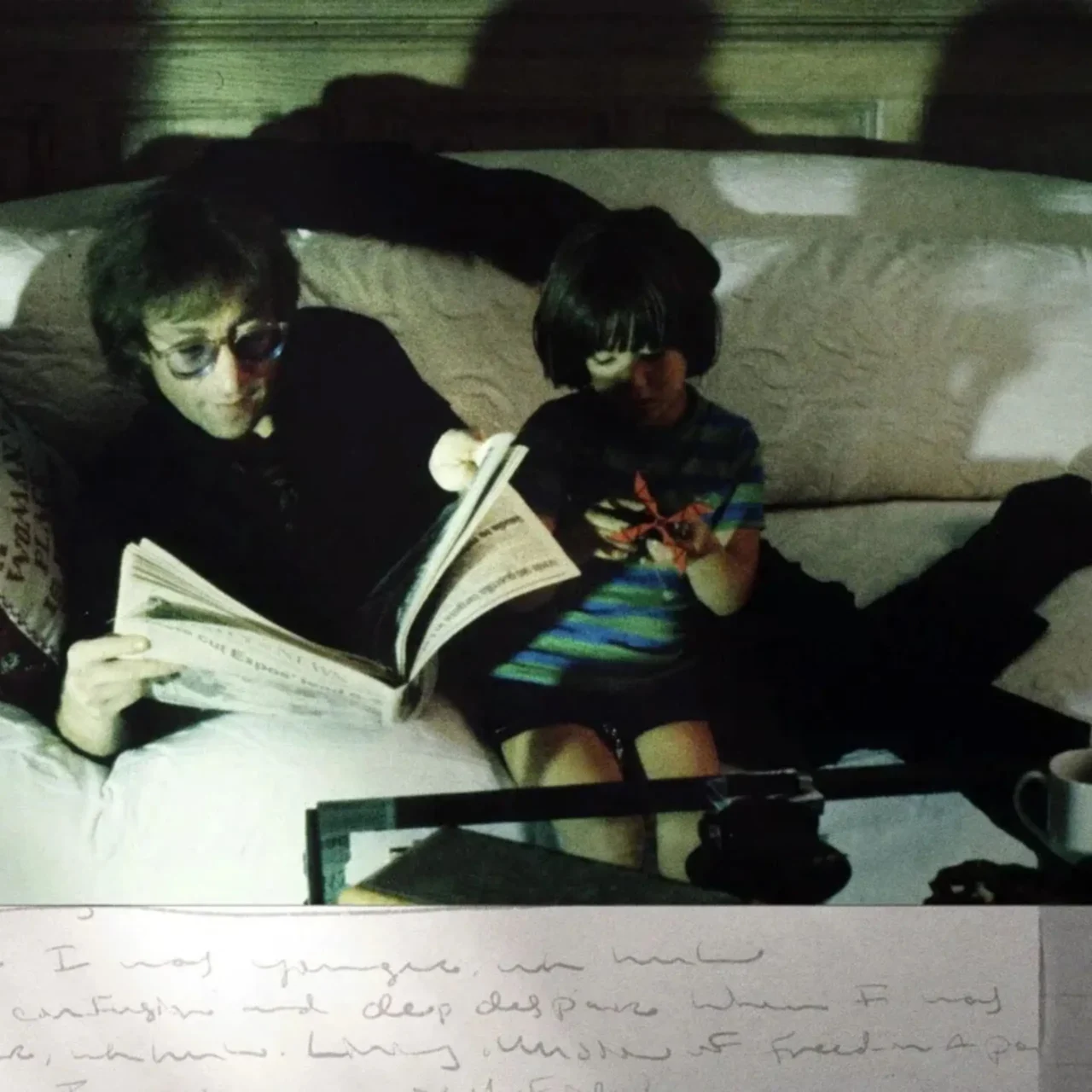 Two people sit closely on a light couch: an adult with glasses reads a newspaper, while a child looks at a small device beside them in a dim room.
