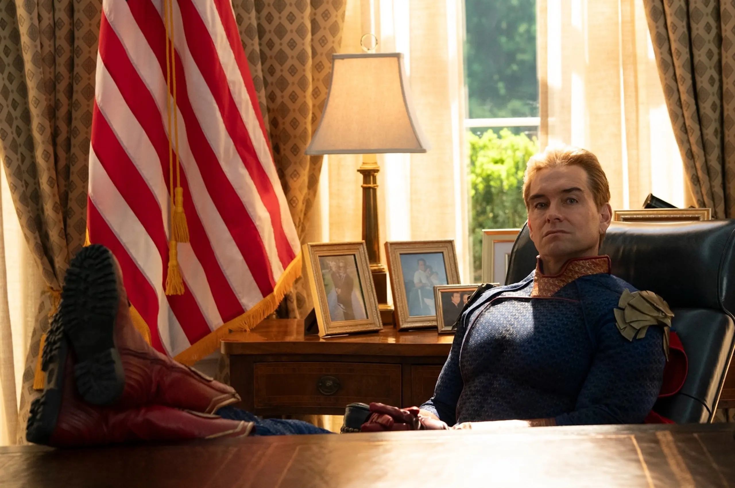 Man in a blue ceremonial uniform sits at a wooden desk with framed photos, a lamp, and an American flag behind him.