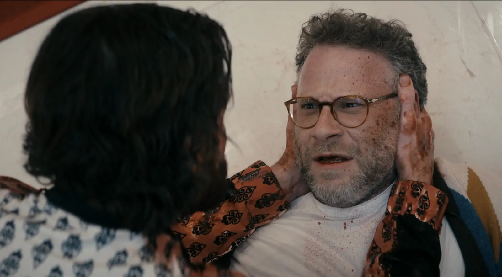 Man with glasses is held by two hands on his cheeks in a tense scene, with blood on his face and shirt in a dim room.
