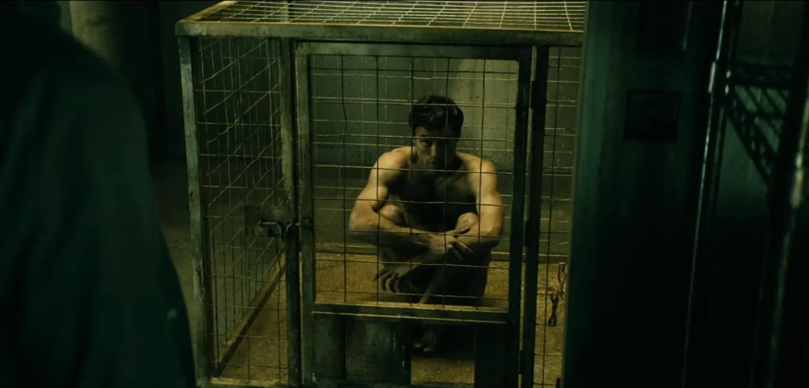 Shirtless man crouches inside a metal cage in a dim, greenish-lit basement-like room.