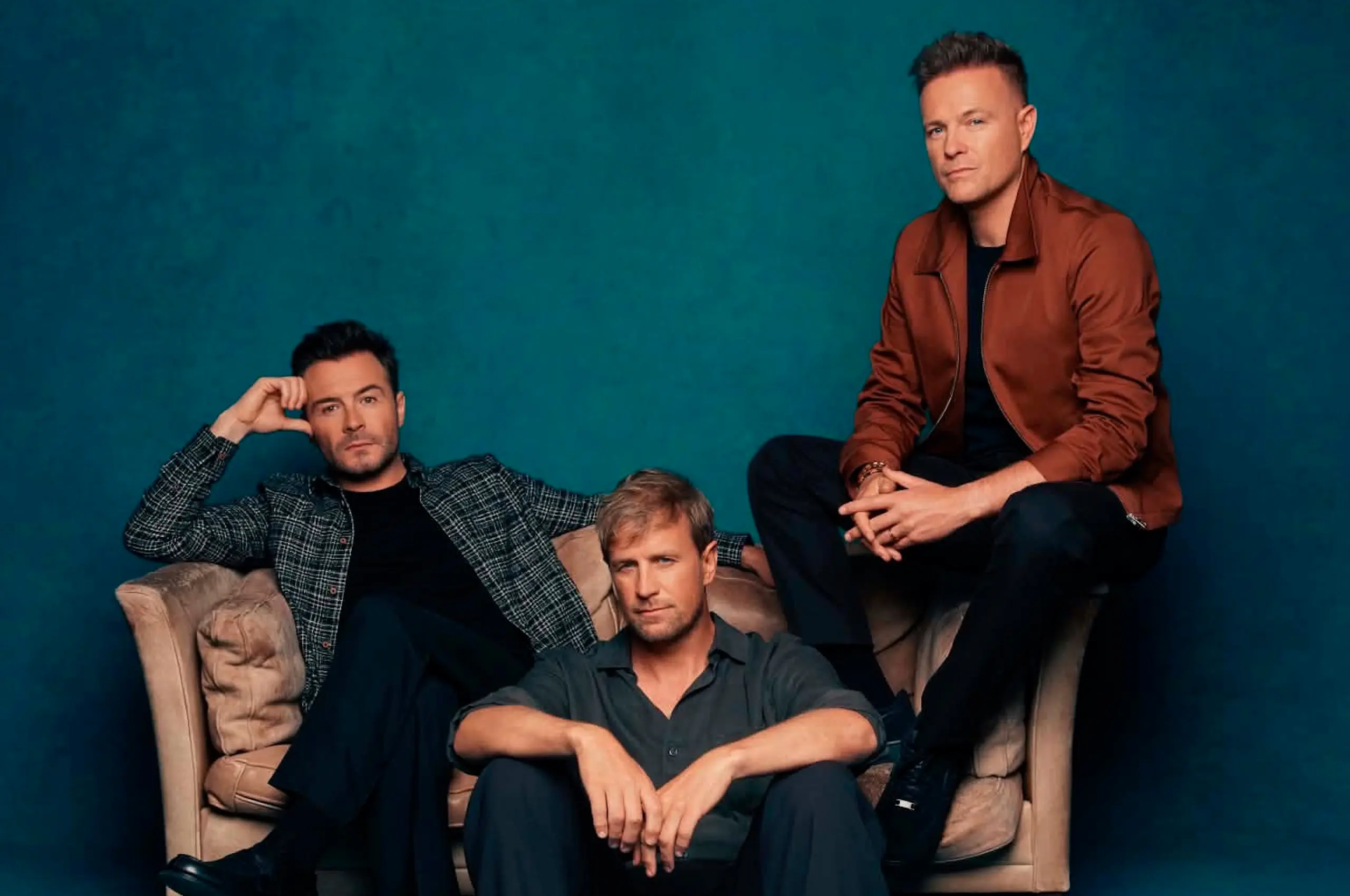 Four men posing on a beige sofa against a teal studio backdrop, wearing casual jackets and jeans westlife