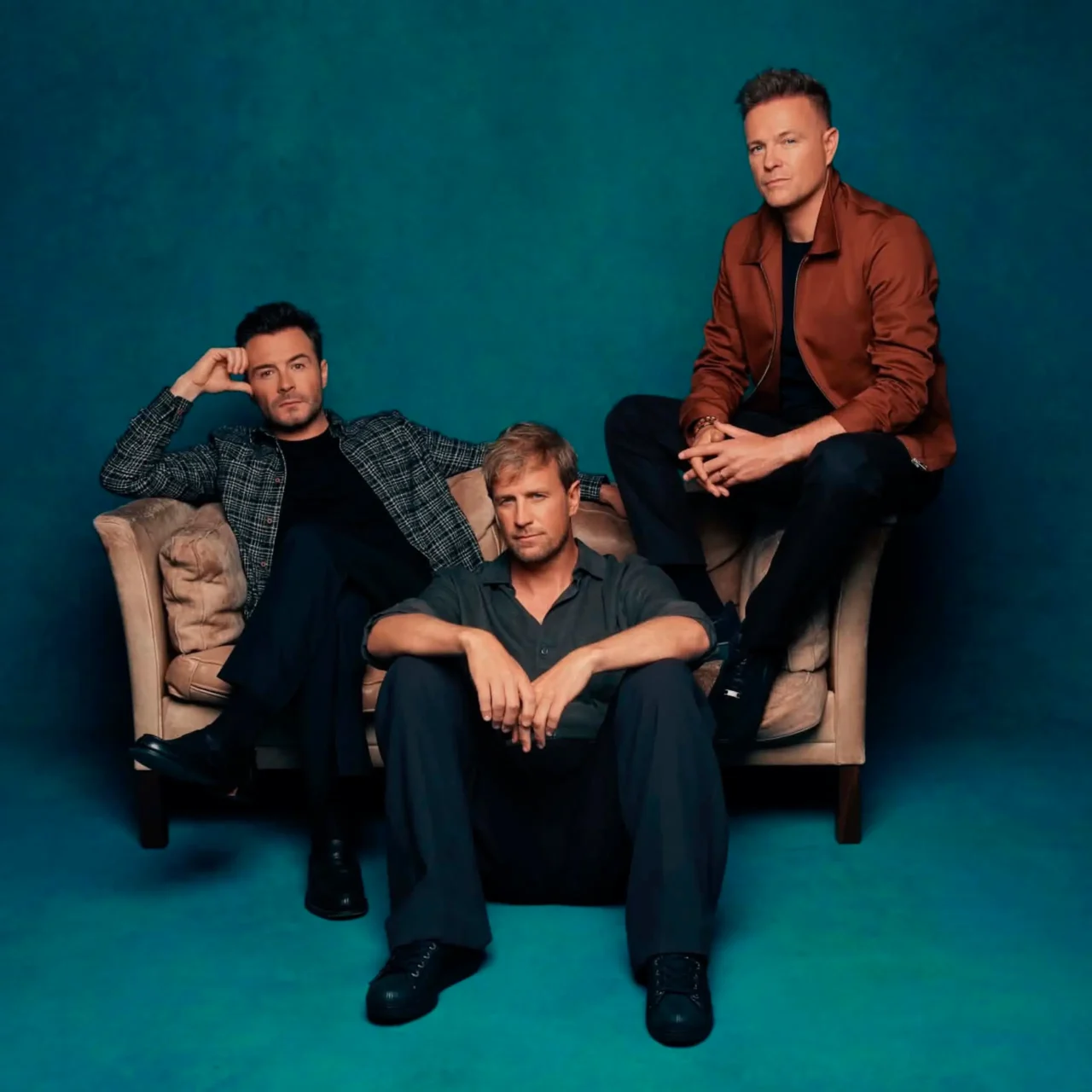 Four men posing on a beige sofa against a teal studio backdrop, wearing casual jackets and jeans westlife